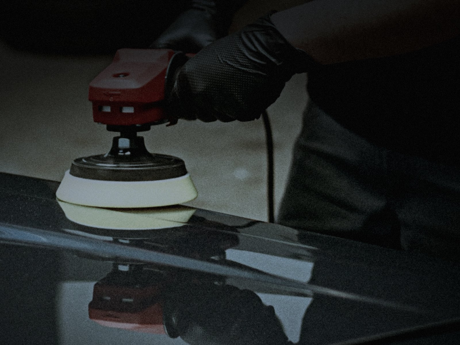 Dual action polisher with a gloved hand working a dark panel in a dim garage